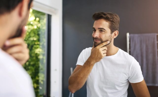 Η περιποίηση της επιδερμίδας, ο καθρέφτης και ο ευτυχισμένος άνθρωπος στο σπίτι με τα χέρια στο πρόσωπο για γενειάδα, επιθεώρηση ανάπτυξης ή τρίχες προσώπου. Μπάνιο, έλεγχο και αρσενικό άτομο με προβληματισμό για καθαρισμό, λάμψη ομορφιάς και αποτελέσματα περιποίησης