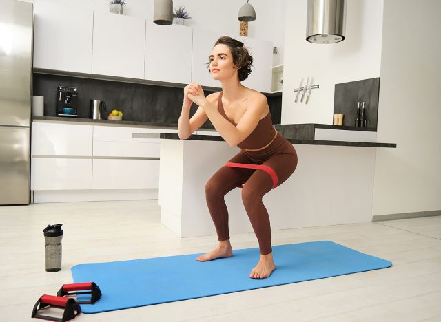Γυμναστήριο στο σπίτι. Young Sportswoman, κορίτσι σε activewear, που κάνει καταλήψεις στην κουζίνα της, προπόνηση σε εσωτερικούς χώρους σε καουτσούκ χαλάκι, χρησιμοποιώντας ελαστική λαστιχένια ζώνη για ασκήσεις.