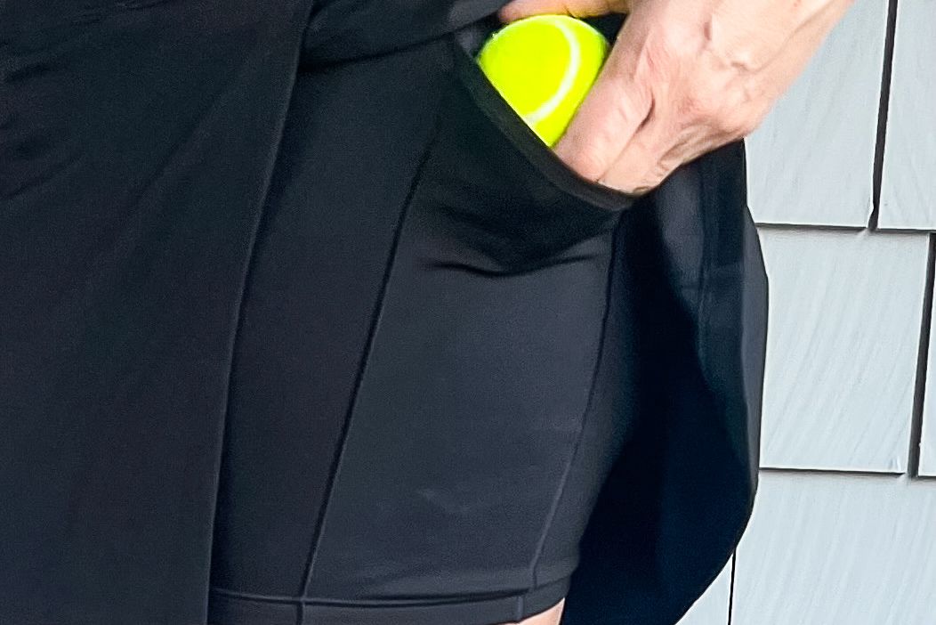 Ένα άτομο που τοποθετεί μια μπάλα τένις στην τσέπη του Athleta Crosscourt Skort